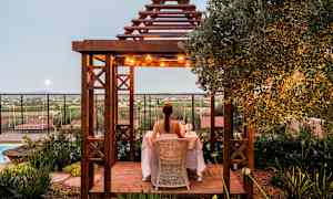 En person sitter vid ett bord under en träpergola som är dekorerad med ljus, omgiven av grönska med utsikt över landskapet. (AI-genererad text)