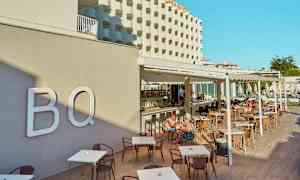 Hotellfasad och utomhusservering med bord och stolar, människor som sitter och äter. (AI-genererad text)