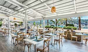 Uteservering vid havet med bord och stolar samt utsikt över båtar i hamnen. (AI-genererad text)