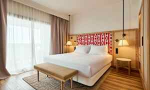 Ett hotellrum med en dubbelsäng, ljusa gardiner och en bänk framför sängen. (AI-genererad text)