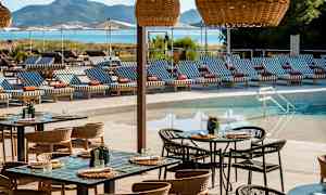 En terrass med bord och stolar framför en pool och solsängar med randiga dynor, med en bergig bakgrund. (AI-genererad text)
