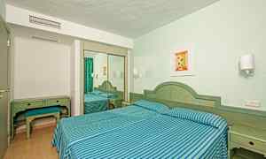 Ett hotellrum med två enkelsängar klädda i blå- och grönrandiga sängkläder, skrivbord med pall och en spegelgarderob. (AI-genererad text)