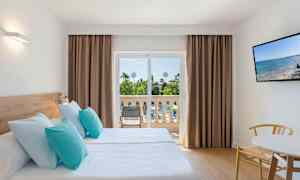 Ett hotellrum med två enkelsängar med ljusblå och turkosa kuddar, en balkong med utsikt över trädgård, och en TV på väggen. (AI-genererad text)