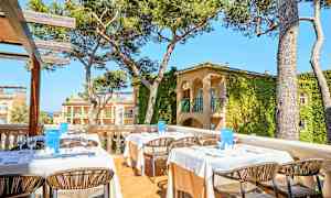 Bild på en utomhusterrass med dukade bord och stolar, omgiven av träd och byggnader med växtbeklädda fasader. (AI-genererad text)