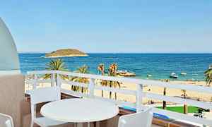 Utsikt från en balkong med ett vitt bord och två stolar mot en sandstrand, palmer och ett blått hav. (AI-genererad text)