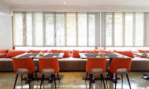 Ett rum med bord och stolar, dekorerat med orange kuddar och solskyddade fönster. (AI-genererad text)