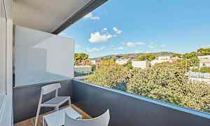 En balkong med två stolar och ett bord, med utsikt över omgivande träd och byggnader under en blå himmel. (AI-genererad text)