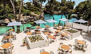 En terrass med bord, stolar och parasoller nära en turkos vattenstrand och grönska. (AI-genererad text)