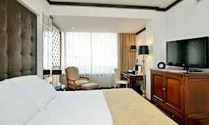 Ett hotellrum med en säng med vitt och beige överkast, en fåtölj och fotpall vid fönstret, ett skrivbord med stol och en TV placerad på en träbyrå. (AI-genererad text)