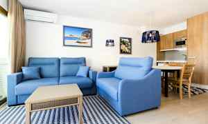 Ett vardagsrum med blå soffor, ett soffbord, vägghängda tavlor och ett kök med trädetaljer synligt i bakgrunden. (AI-genererad text)