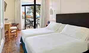 Ett ljust hotellrum med två enkelsängar, ett skrivbord och en balkong med utsikt över havet och palmer. (AI-genererad text)