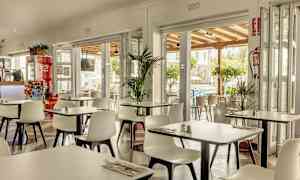 En restaurang med vita stolar och bord, öppen mot en terrass och omgivande grönska. (AI-genererad text)