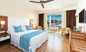 Ett hotellrum med en stor säng, blå dekorativa kuddar och täcke, samt ett bord med stolar vid ett glasfönster som leder till en balkong. (AI-genererad text)