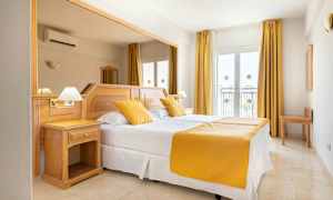 Ett hotellrum med en dubbelsäng, vita lakan och gula kuddar och överkast. Rummet har ljust trämöblemang och fönster med gardiner. (AI-genererad text)