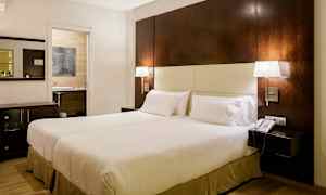 Ett hotellrum med en stor säng, två sänglampor, ett nattduksbord och en telefon. En öppning mot ett badrum syns i bakgrunden. (AI-genererad text)