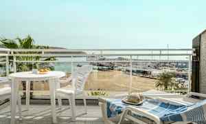 Vy från en balkong med vita utemöbler, med utsikt över en strand, en småbåtshamn och havet i bakgrunden. (AI-genererad text)