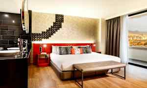 Ett hotellrum med en stor säng, modern inredning och en sittbänk framför sängen. Ett fönster visar utsikten mot berg och byggnader. (AI-genererad text)