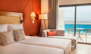 Ett hotellrum med dubbelsäng, dekorativa kuddar, en soffa och glasdörrar som leder till balkongen med utsikt över havet. (AI-genererad text)