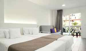 Ett hotellrum med två enkelsängar med vita sängkläder, en grå soffa med färgade kuddar och en balkong med utsikt. (AI-genererad text)