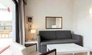 Ett vardagsrum med en grå soffa och ett vitt bord, en fåtölj och en fönsterdörr till en balkong. (AI-genererad text)