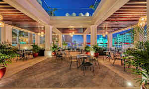 En uteservering med bord och stolar under en takkonstruktion, omgiven av palmer och belysning. (AI-genererad text)