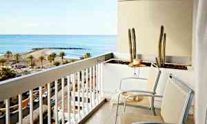 En balkong med två stolar och en blomlåda med kaktusar. Utsikten visar en strand med palmer och en havsvik. (AI-genererad text)