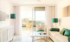 Ljust vardagsrum med en soffa, bord och stolar samt glasdörrar som leder till en balkong med utsikt över havet. (AI-genererad text)