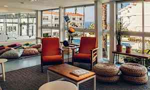 Ett ljust rum med stora fönsterdörrar, två orangea fåtöljer och korgpallar framför ett kaffebord. (AI-genererad text)