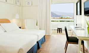 Ett ljus och modernt hotellrum med två enkelsängar, en vit fåtölj och ett skrivbord med lampa. Ett stort fönster leder ut till en balkong med utsikt över berg och grönska. (AI-genererad text)