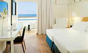 Ett hotellrum med två enkelsängar, skrivbord med stol, en vit fåtölj och utsikt mot havet från en privat balkong. (AI-genererad text)