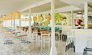 Ett öppet uteområde med restaurangmöbler och ljusa inredningsdetaljer, såsom vita bord och stolar samt korgar. Dekorativa lampor hänger från taket. (AI-genererad text)