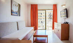 Vardagsrum i en semesterlägenhet med en soffa, ett soffbord, en byrå med en TV och en balkong med utemöbler. (AI-genererad text)