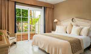 Ett hotellrum med en stor säng, beige sängkläder, vita kuddar och en glasdörr som öppnar mot en terrass med grönska. (AI-genererad text)