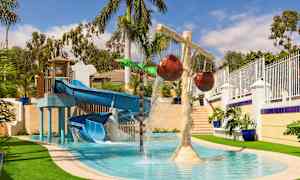 En vattenlekplats med en blå rutschkana och vattenstrålar samt dekorativa palmer. (AI-genererad text)