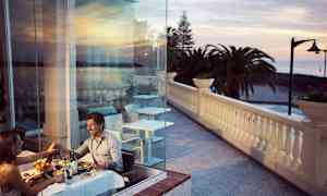 Par som äter middag vid ett bord nära glasvägg med utsikt över terrass och hav. (AI-genererad text)