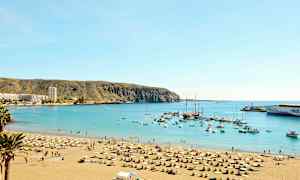 Strand med solstolar och parasoller vid en skyddad havsbukt med båtar och byggnader synliga i bakgrunden. (AI-genererad text)