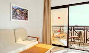 Utsikt från ett hotellrum med en beige soffa, ett träbord och en balkong med utsikt över stranden. (AI-genererad text)