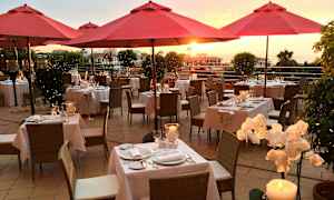 Utomhusrestaurang med röda parasoller, dukade bord och solnedgång i bakgrunden. (AI-genererad text)