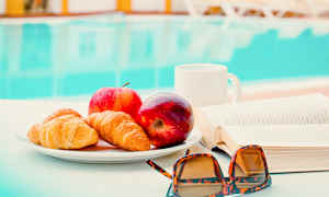 En tallrik med croissanter och äpplen står på ett bord bredvid en bok och ett par solglasögon. I bakgrunden syns en pool. (AI-genererad text)