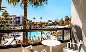 Utsikt från en balkong över ett hotellområde med pool och solstolar, omgiven av palmer. (AI-genererad text)