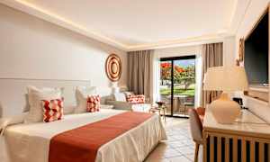 Ett ljust och elegant hotellrum med en stor säng, dekorerade kuddar, och en privat sittplats utomhus med vy över en trädgård. (AI-genererad text)