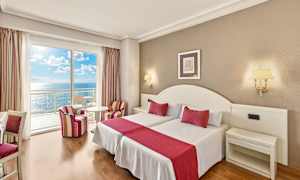 Ett hotellrum med två bäddar, röda accenter och havsutsikt från en balkong. (AI-genererad text)