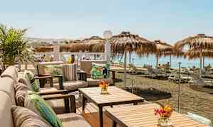 Uteplats med sittmöbler och bord med utsikt över en strand med parasoller och solstolar. (AI-genererad text)