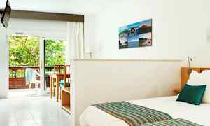 Ljus hotellrum med säng, bord och stolar samt utsikt mot en grönskande balkong. (AI-genererad text)