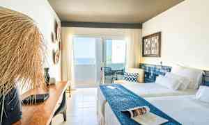 Ett ljust hotellrum med två bäddar, en träbänk och en balkong med havsutsikt. (AI-genererad text)