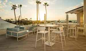 Utsikt över en möblerad terrass med bord och stolar vid solnedgången, med palmer och havet i bakgrunden. (AI-genererad text)