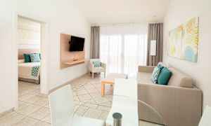 Bild föreställer ett ljust och modernt vardagsrum med en soffa, TV samt matbord och stolar. En dörr leder till ett sovrum. (AI-genererad text)