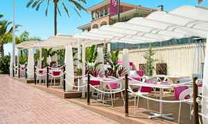 Utomhusservering med vita parasoller och rosa dukar på bord. Palmer och en byggnad i bakgrunden. (AI-genererad text)