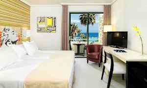 Ett hotellrum med en säng, en stol, ett skrivbord och en TV. Utanför ett glasfönster syns en balkong med havsutsikt. (AI-genererad text)