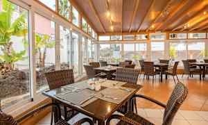 En matsal med rottingstolar och bord i trä, omgiven av stora glasfönster med utsikt mot en trädgård. (AI-genererad text)